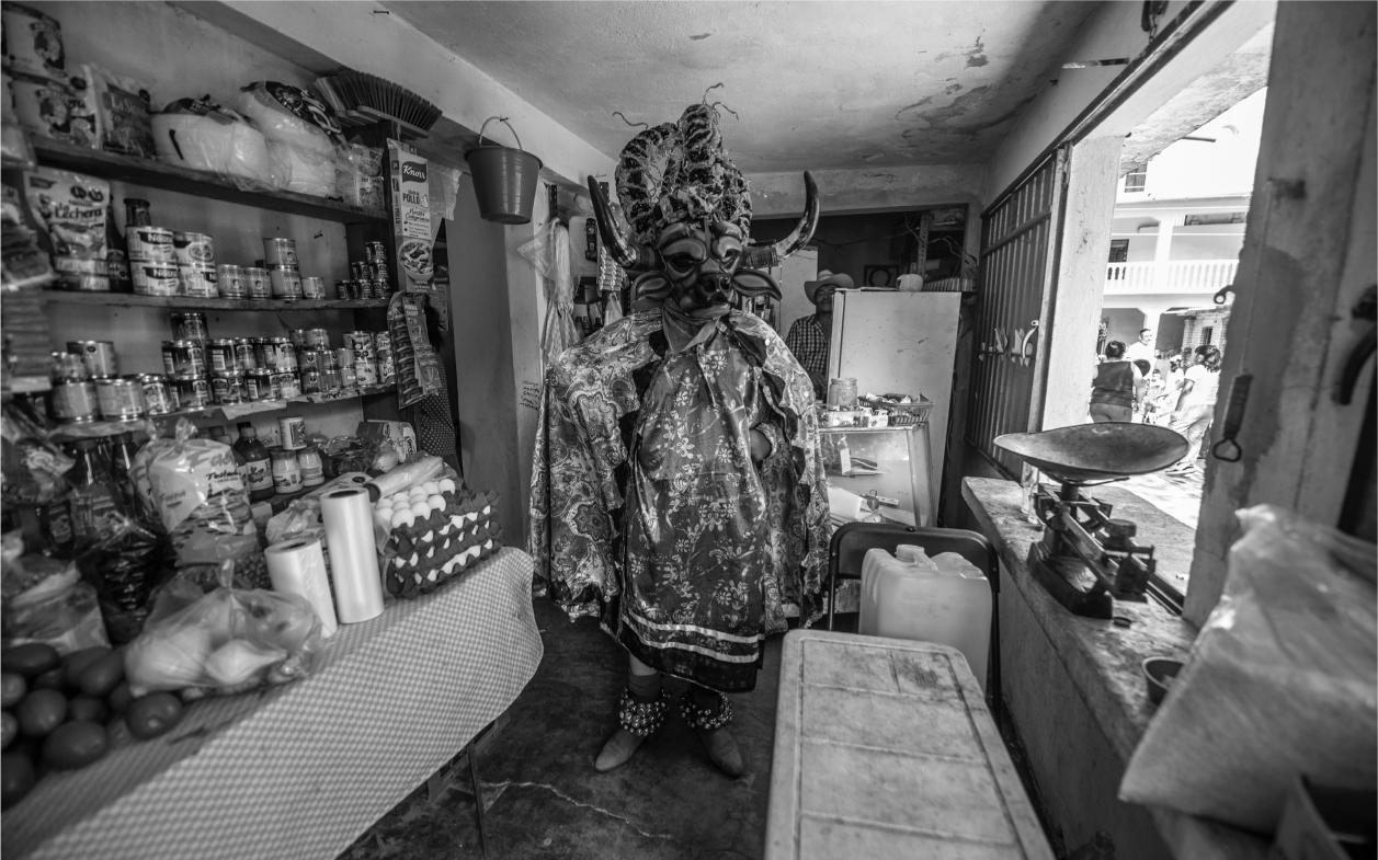 A photograph of an Afro-Mexican ceremony in Coyolillo, Mexico. A person dressed in an ornate bull’s head mask and embroidered cloak stands in a small grocery or general store. Foodstuffs are stacked on tables and shelves to the left, and a scale is visible on the right in front of a window that opens upon the street.
