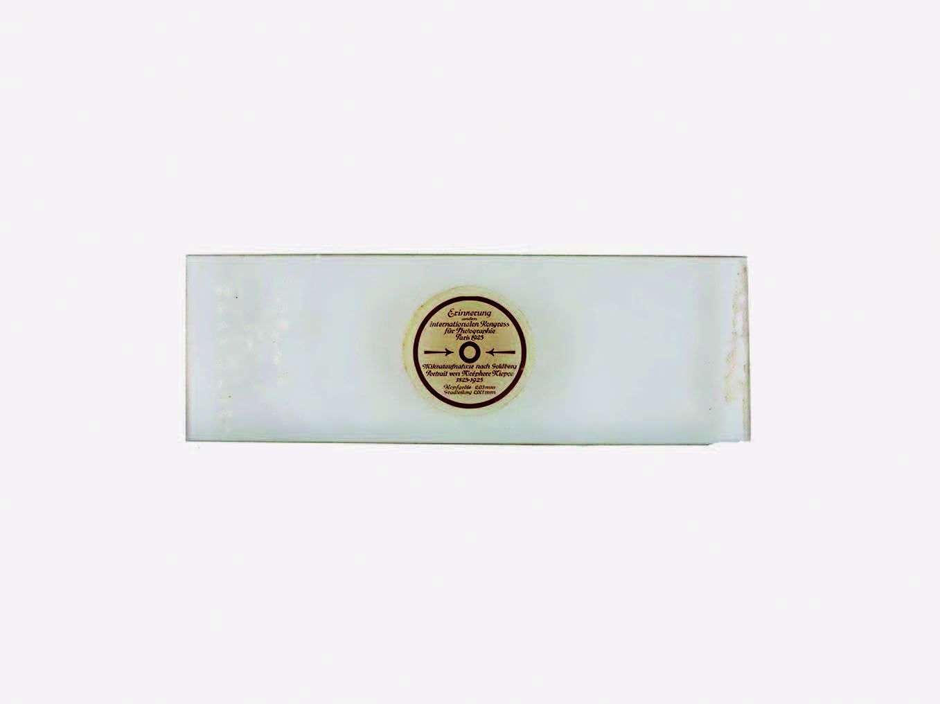 A slide of white glass, featuring a paper decal in the shape of a circle, with a note in German, stating that the slide contains a work of microscopic portraiture depicting Nicéphore Niepce.