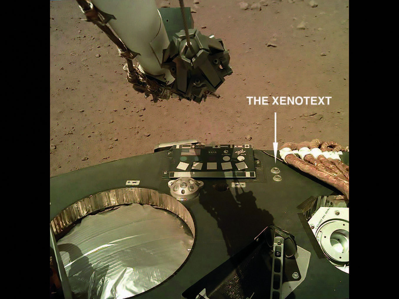 A photograph showing a robot scoop poised above the deck of the InSight lander on the russet, barren surface of Mars, complete with an array of intrumentation aboard the probe. The photo is labelled with text reading ‘The Xenotext’ and an arrow indicating the position on the probe of a microchip containing this poem.