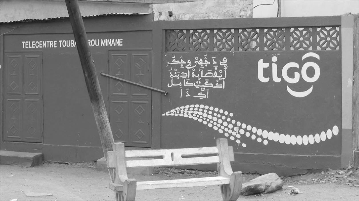 A photograph of a painted wall that highlights the use of A’jami in telecommunication centers. Text and a decorative motif is painted on the wall in white. There is a bench and telephone pole visible in the foreground.