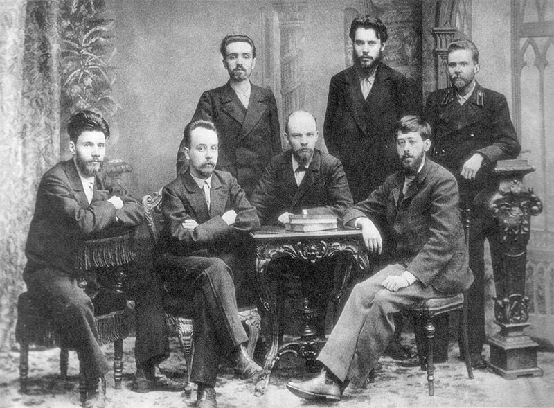 A photograph of Lenin, Martov, and other young intellectuals posing for a picture. Lenin sits at a table in the center with Martov seated to his left. Two other men are seated to Lenin's right, and three men stand behind him.