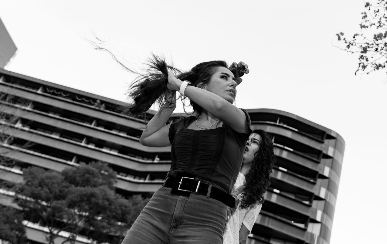 A photograph of a woman having her hair shaved off in solidarity with Iranian protests for women’s rights. A second woman stands behind her holding electric clippers to her head. There is an apartment building visible in the background behind the women.