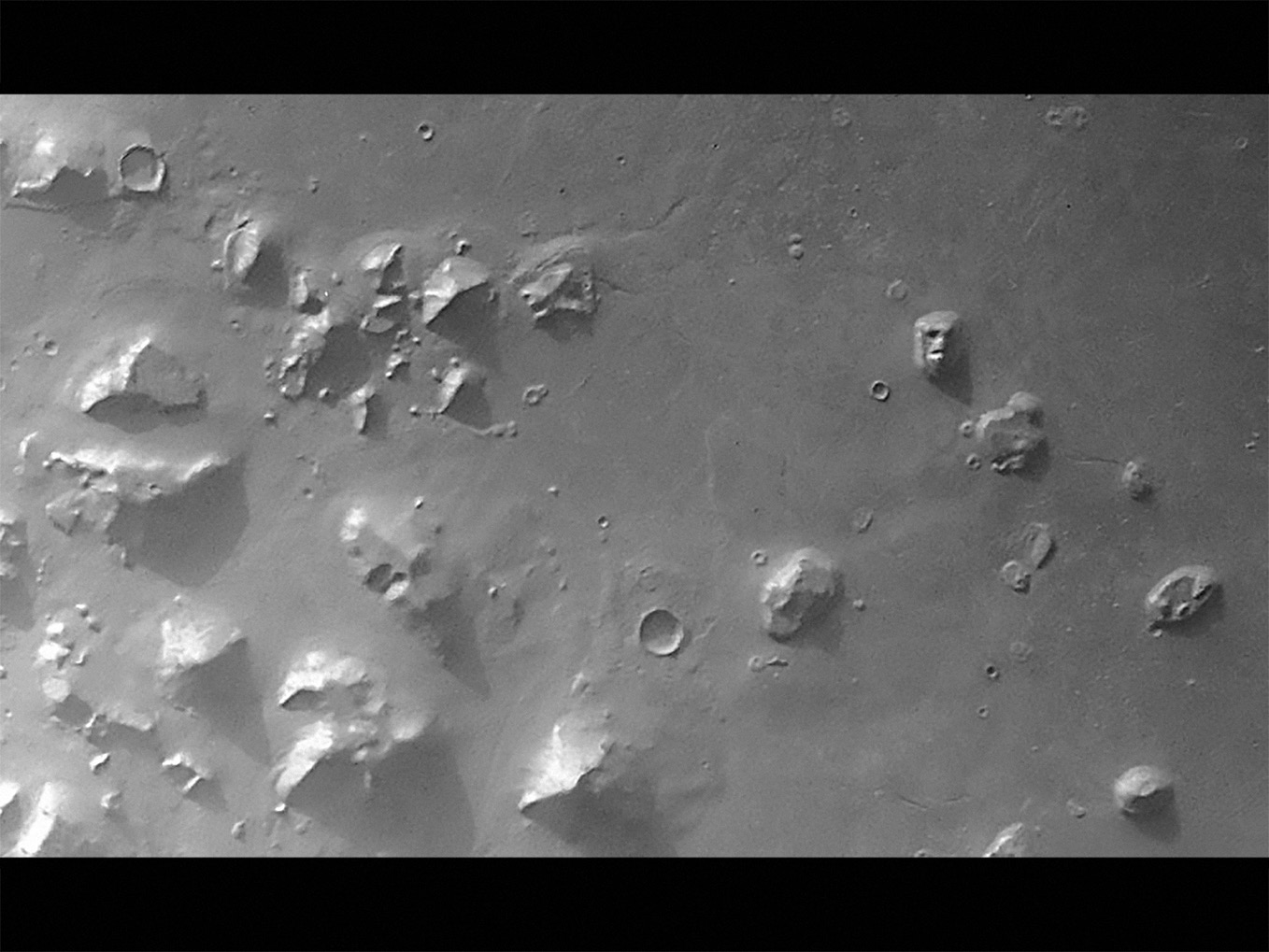 A monochrome photograph, showing an aerial image of the Cydonia Planitia on Mars, complete with massifs and craters, including the purported monument of a ‘face,’ carved into a mountain.